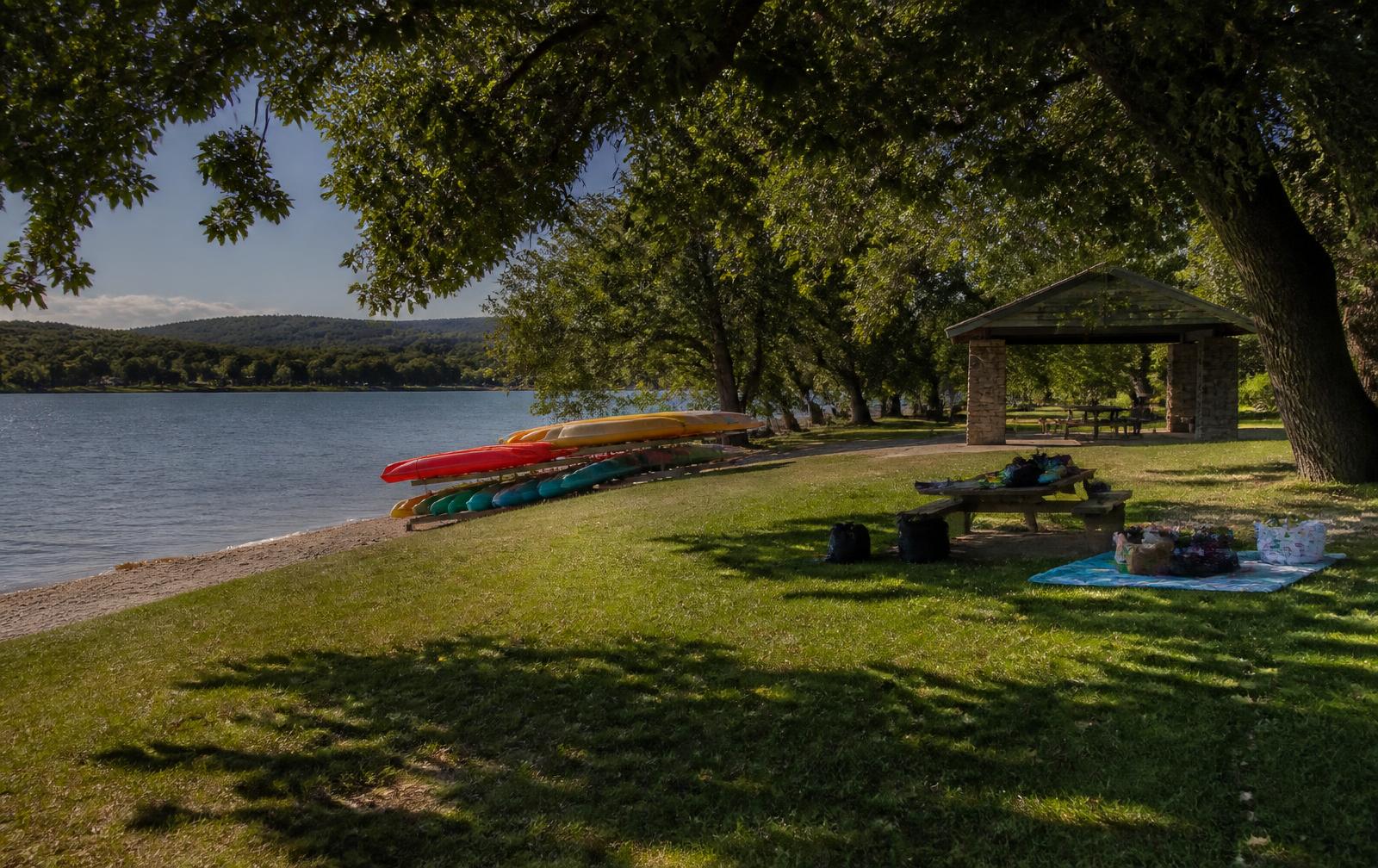 Shaded lakeside park with kayak racks, a picnic pavilion, and a small sand beach
